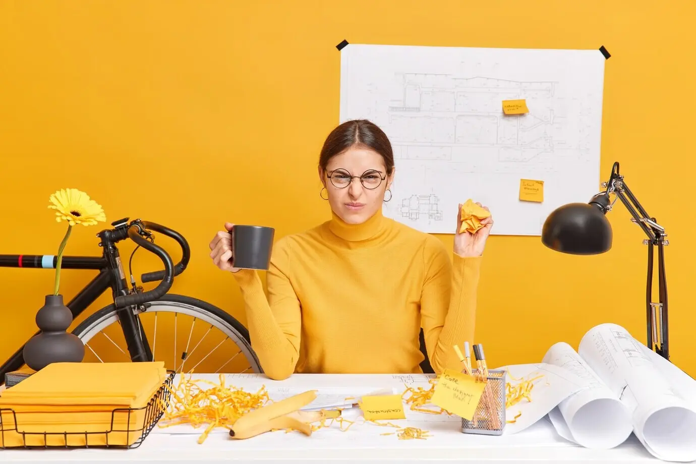An unhappy female architect frowns, dissatisfied with her architectural project; she crumples papers, drinks coffee, wears spectacles and a turtleneck, and sits at a desk.