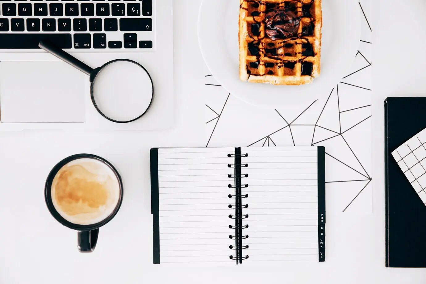 On a white desk: a coffee cup, a laptop, magnifying glasses, a spiral notepad, and a plate of waffles with chocolate.